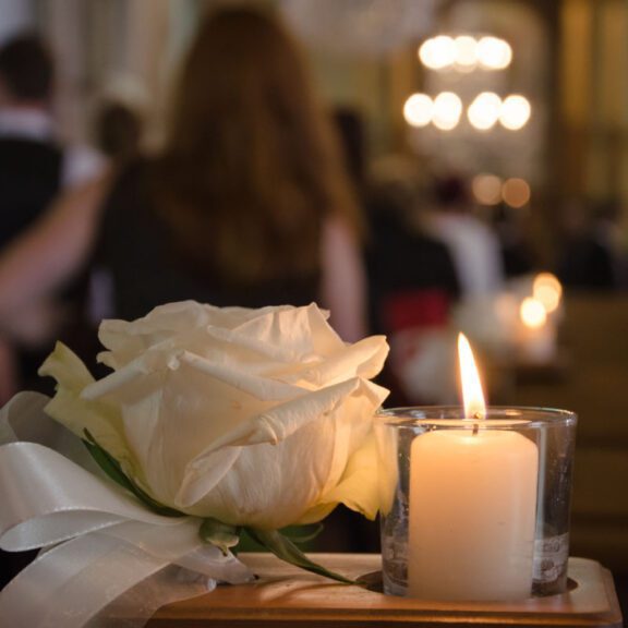 photo of a white rose with a ribbon attached to the bottom next to a white candle burning in a clear candle holder